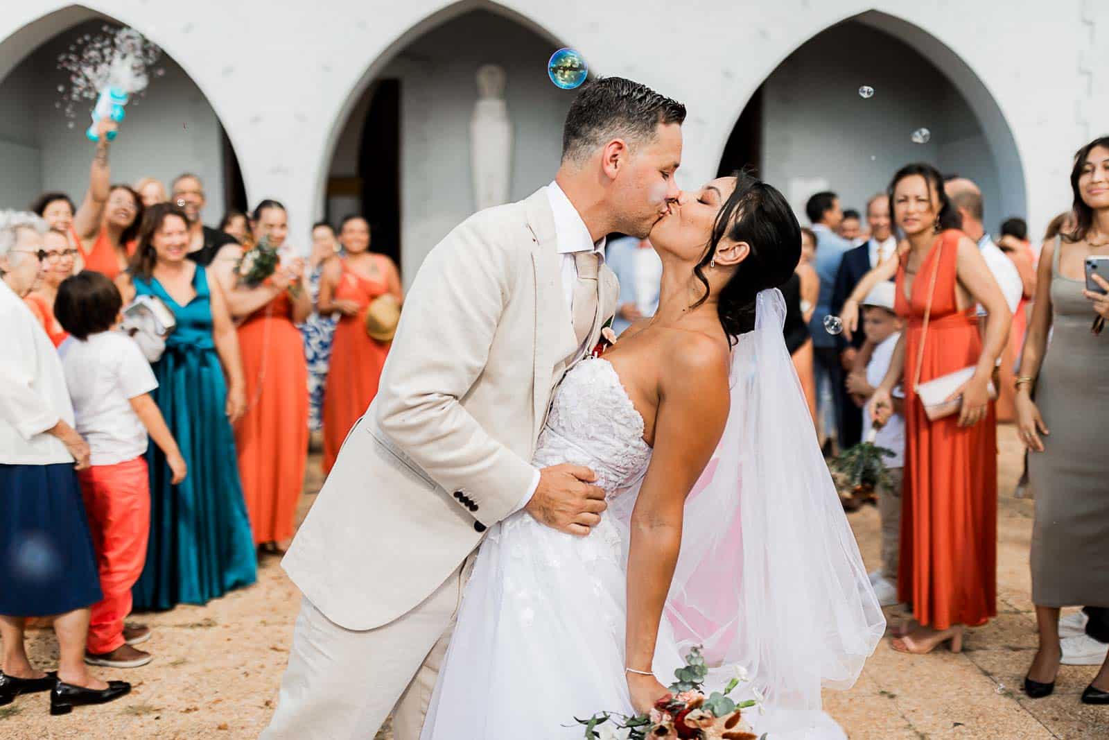 Photographie de Mathieu Dété, photographe de mariage et famille à Saint-Pierre sur l'île de la Réunion 974, présentant le bisou des mariés à la sortie de l'église de Saint-Gilles, devant leurs invités